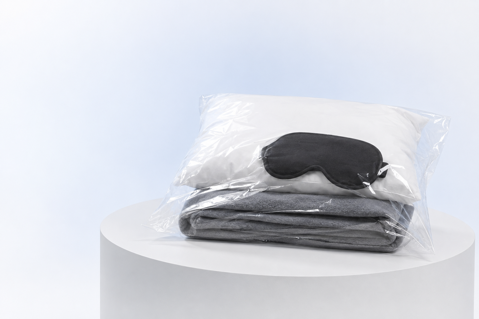 Kit de viaje con almohada blanca, manta gris y antifaz negro dentro de una bolsa plástica transparente